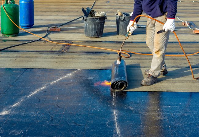 Worker preparing part of bitumen roofing felt roll for melting by gas heater torch flame
