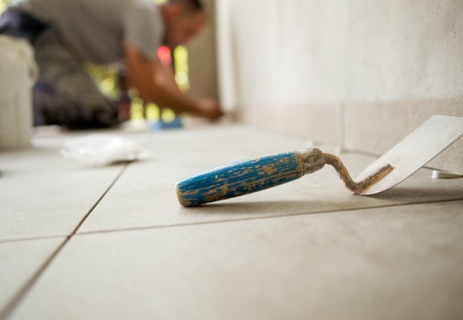 Trowel is on the floor and behind is a worker gluing ceramic tiles.